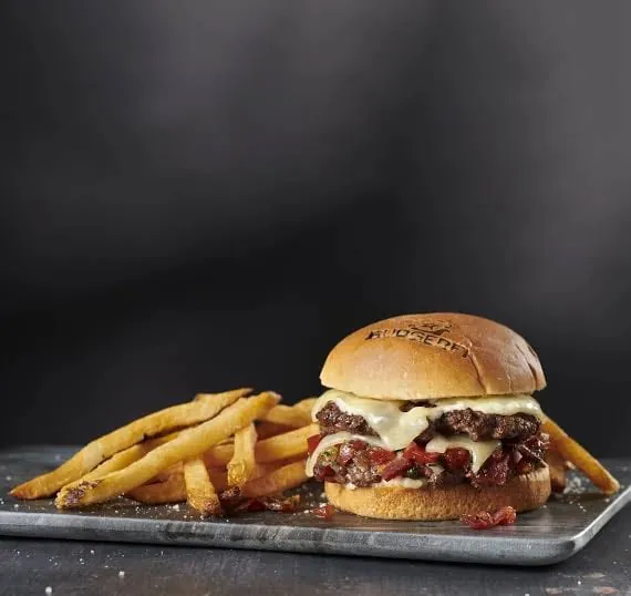 Hamburguesa artesanal con queso fundido y papas fritas, servida en una bandeja de metal.