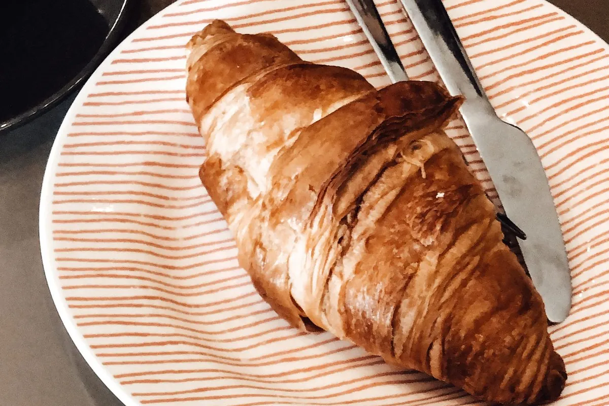 Croissant recién horneado sobre un plato de rayas, junto a una taza de café.